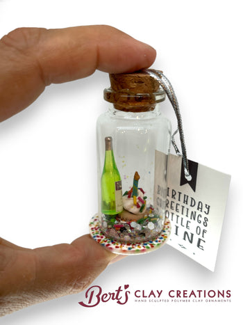 Miniature bottle with a cork top, containing a small scene, held by a hand with a 'Bert's Clay Creations' tag.